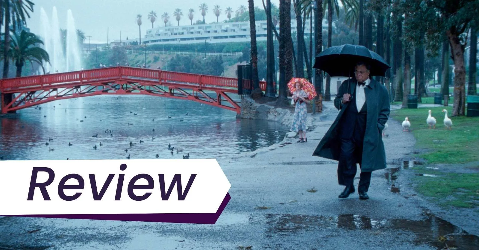 James Earl Jones and Lynn Redgrave star in Charles Burnett's The Annihilation of Fish. Photo courtesy of Milestone Films/Kino Lorber. On a rainy day, an elderly Black men dressed in a black suit with a black umbrella walks by the water, followed by a white woman wearing a patterned dress with a red umbrella. They are walking in a park by the water, surrounded by palm trees and a red bridge.