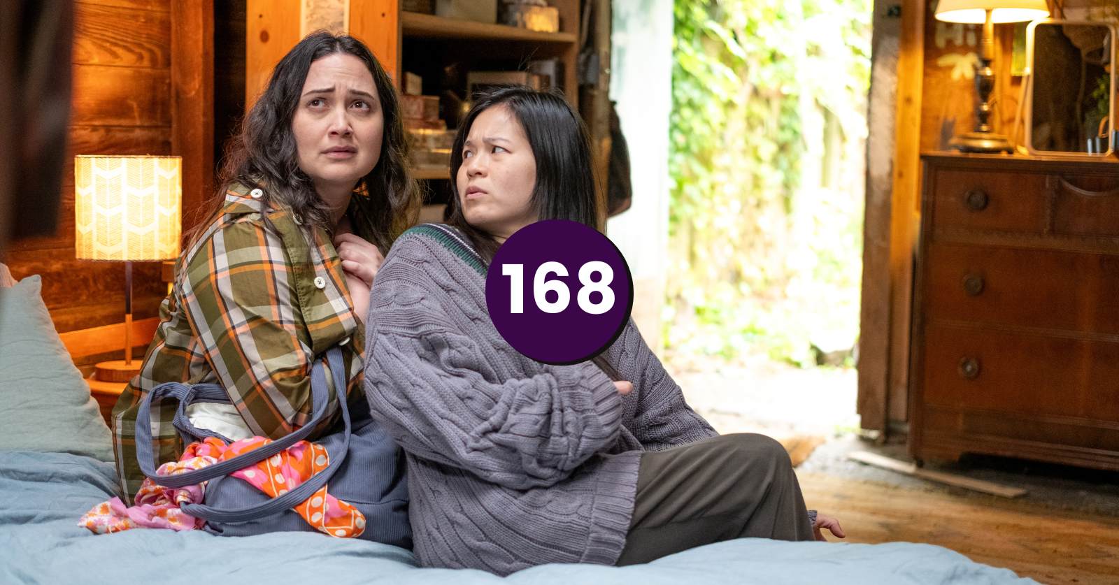 Still from Andrew Ahn's new film The Wedding Banquet which we recommend on the podcast. An Indigenous woman with long brown hair and a Chinese woman sit on a bed in a wood cabin.