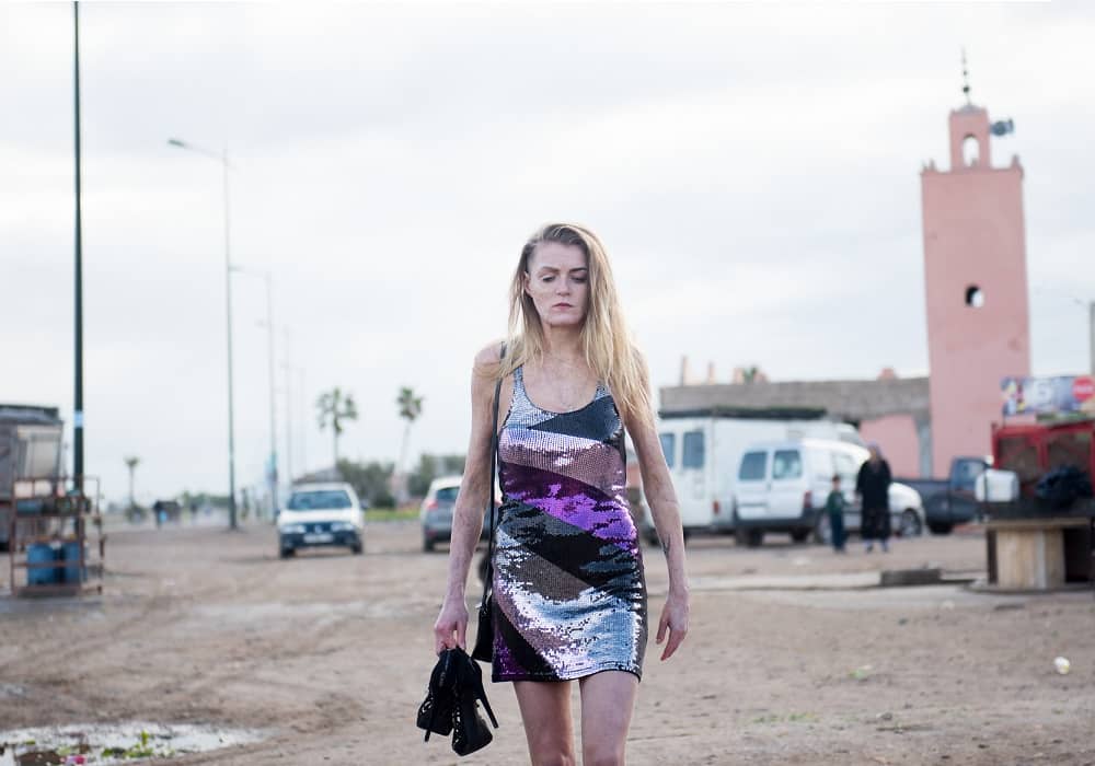 A scarred woman (Vicky Knight) wearing a dress walks through a parking lot in a still from Dirty God
