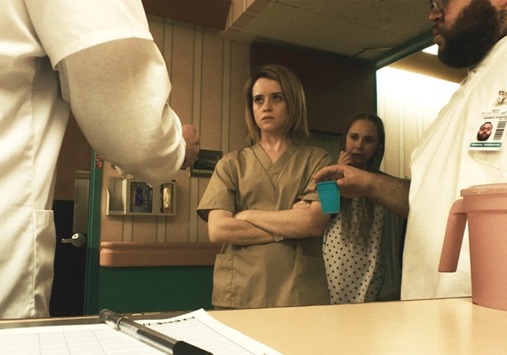 Claire Foy stars in Unsane, Steven Soderbergh. She is wearing hospital scrubs and being served medication. Her arms are crossed and she looks angry.