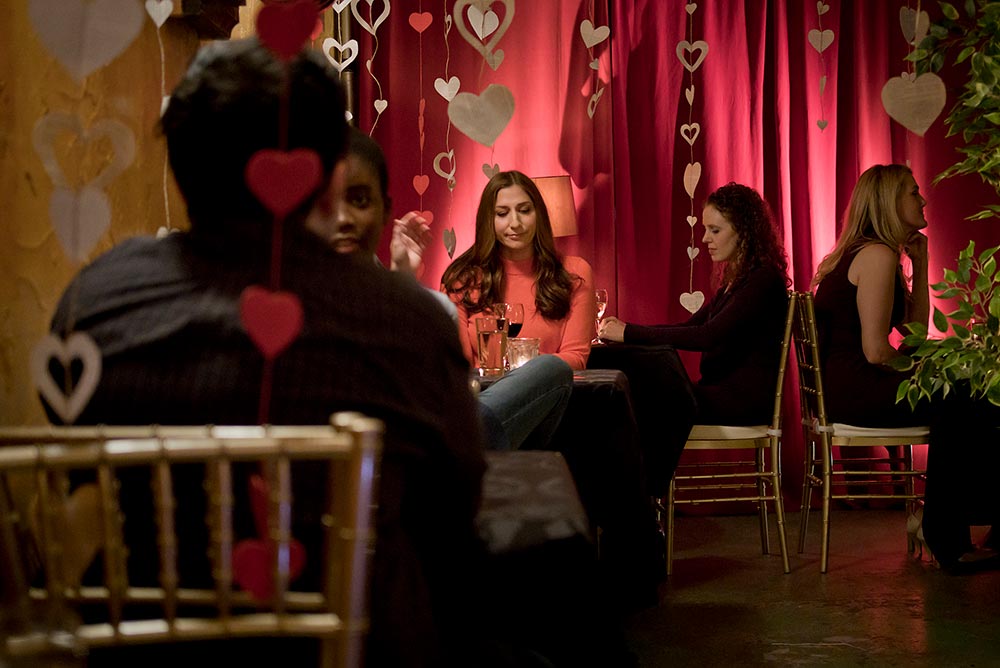 A still of Chelsea Peretti as Gaby, a woman sitting at a table on Valentine's Day, looking sad while she is surrounded by paper cutout hearts and hot pink drapes.