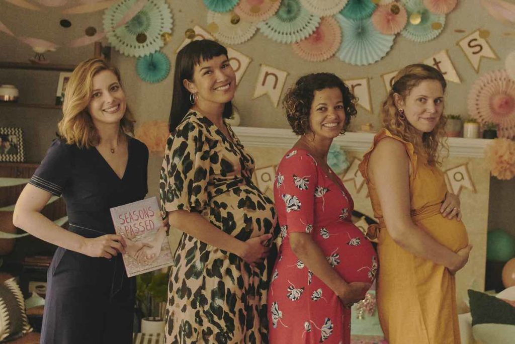 Gillian Jacobs as Kate poses for a photograph in a lineup of pregnant women. She holds her book, Seasons Passed, where her baby bump would be and struggles to smile. Still from I Used to Go Here directed by Kris Rey.