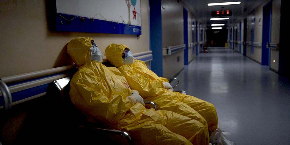 Two figures in PPE suits lie down in a hospital corridor, exhausted.