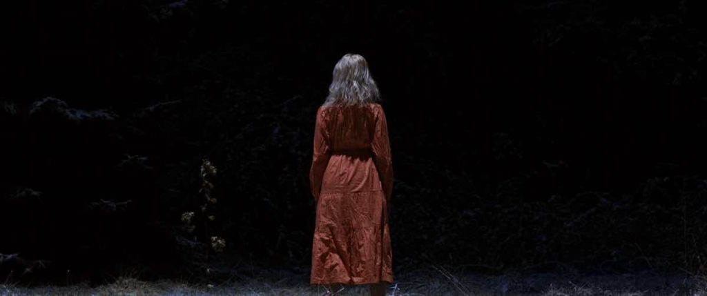 Still from the film Sleep, directed by Michael Venus and screening at Fantasia Festival. A blond woman is centre frame against a mostly black background. She's wearing a red coat, her back turned to the camera.