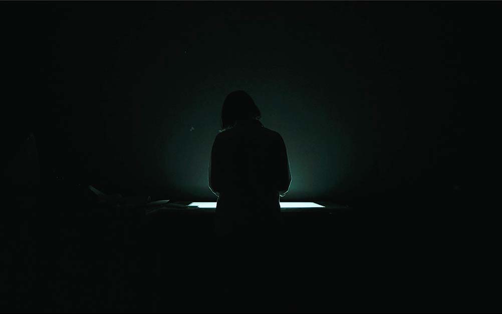 A woman stands in a dark room in front of a film processing device, which illuminates her silhouette.