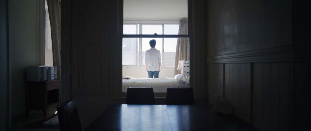 Justin Chon stars as Chang-rae in Wayne Wang's Coming Home Again. Chang-rae is looking out the window at the back of the frame in his mother's bedroom, back turned to camera, seen through a window.