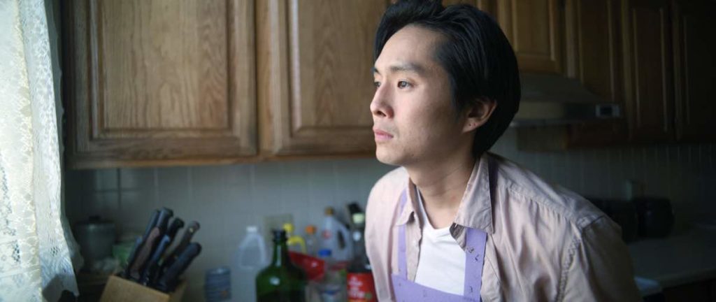 Justin Chon stars as Chang-rae wearing an apron in the kitchen, looking out the window in despair, in Wayne Wang's Coming Home Again.