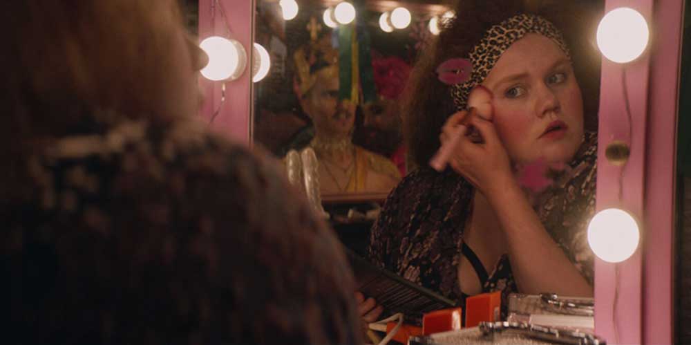 A young woman puts on drag makeup in front of a mirror.
