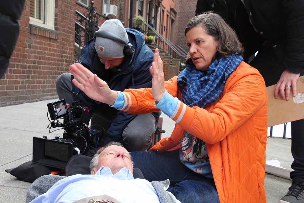 Dick Johnson lies on the street playing dead as filmmaker Kirsten Johnson perches above him, motioning with her arms as a way to direct her crew.