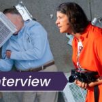 A still from Dick Johnson is dead in which Dick Johnson lies on the pavement after being struck on the head by an air conditioner. In the foreground of the image is director Kirsten Johnson, who is superimposed onto the still. The image also features the text, 'Interview.'