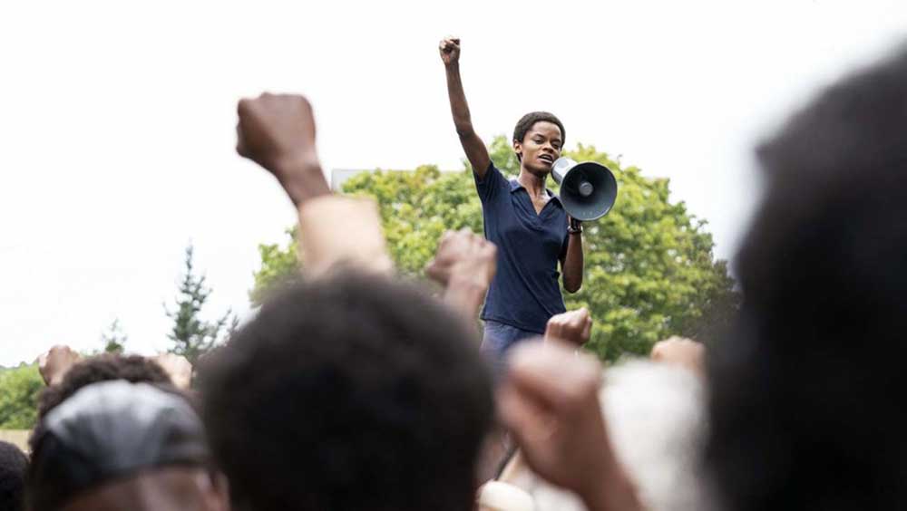 A woman stands on a platform with a a megaphone at a protest, her fist raised in the air.