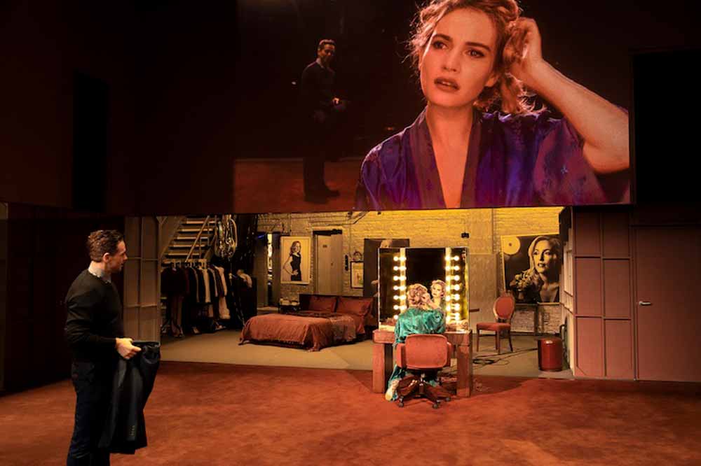Lily James, in the stage production, looks into a mirror on stage while a screen above her projects an image of her face.