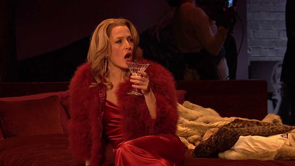 Gillian Anderson as Margo in the 2019 stage production of All About Eve, drinking a martini in a red dress and furs.
