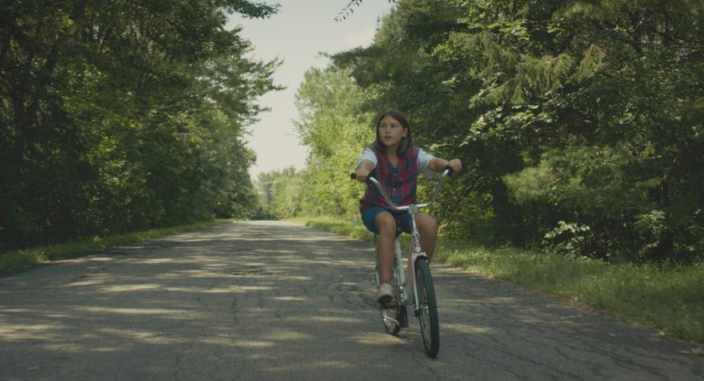 Lake Delisle stars as Ivy in Rustic Oracle, directed by Sonia Boileau. Ivy is riding her bike on a rural road.