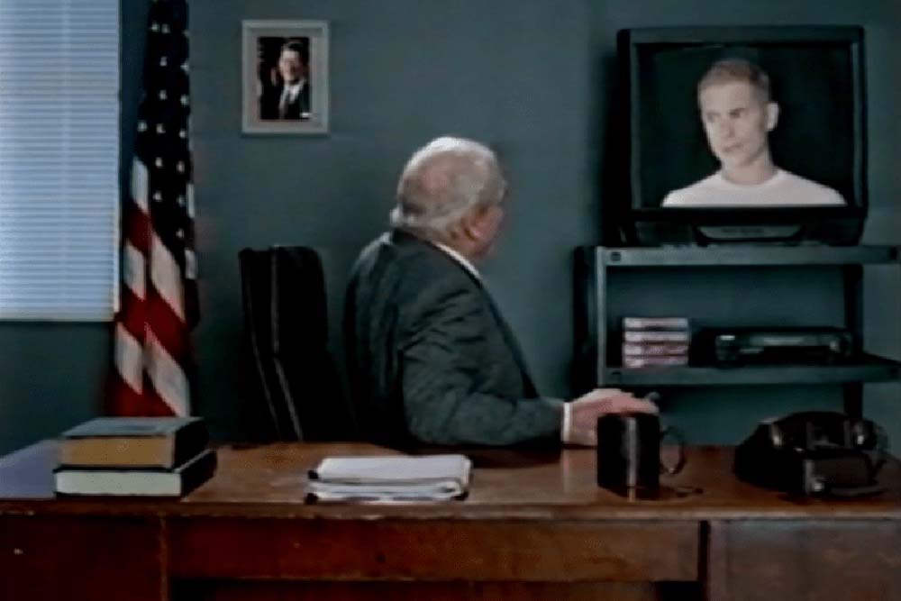 The Narrator turns to look behind him at a small TV set, containing an image of a man dressed in white.