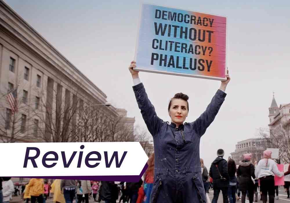 A woman holds up a sign that reads 'Democracy without cliteracy? Phallusy.' The image is a still from The Dilemma of Desire.