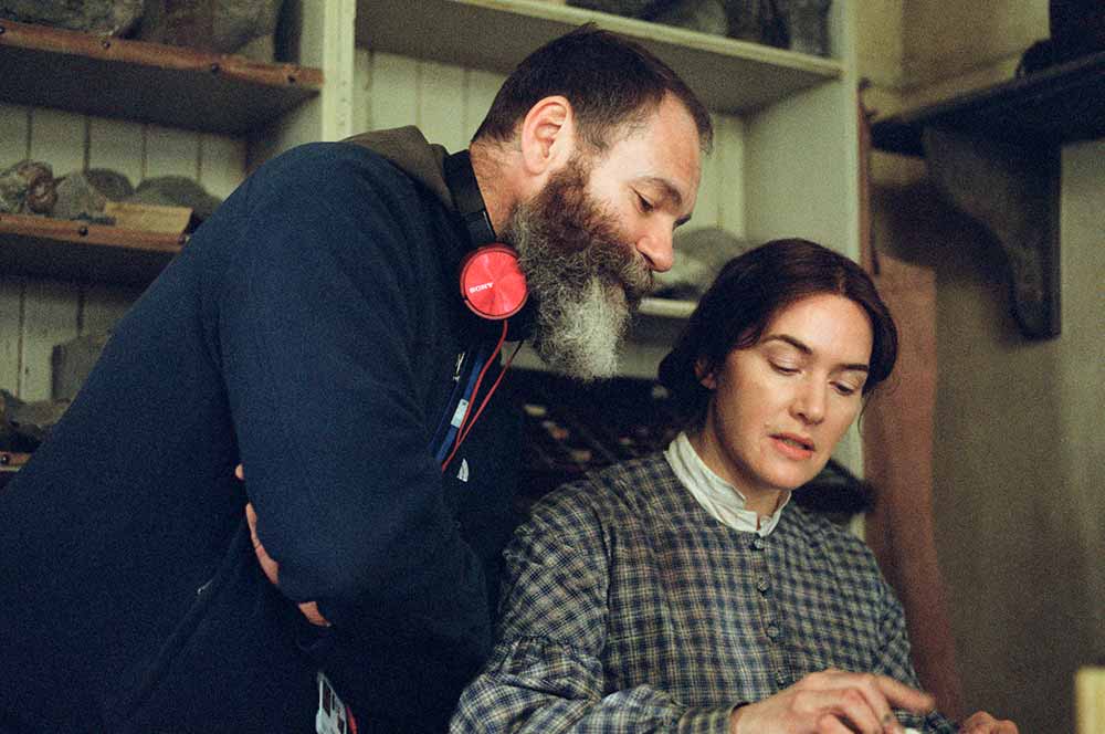 Francis Lee, wearing headphones around his neck, leans over Kate Winslet on the set of Ammonite.