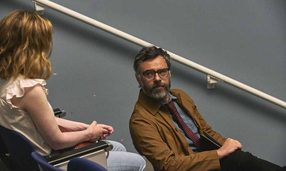 Jemaine Clement sits at a chair in a college lecture hall, looking up at the woman behind him in I Used to Go Here.