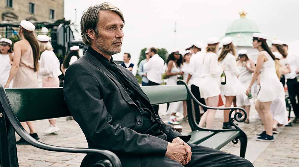 Mads Mikkelsen sits on an outdoor bench in front of a group of teens who have just graduated high school in Another Round.