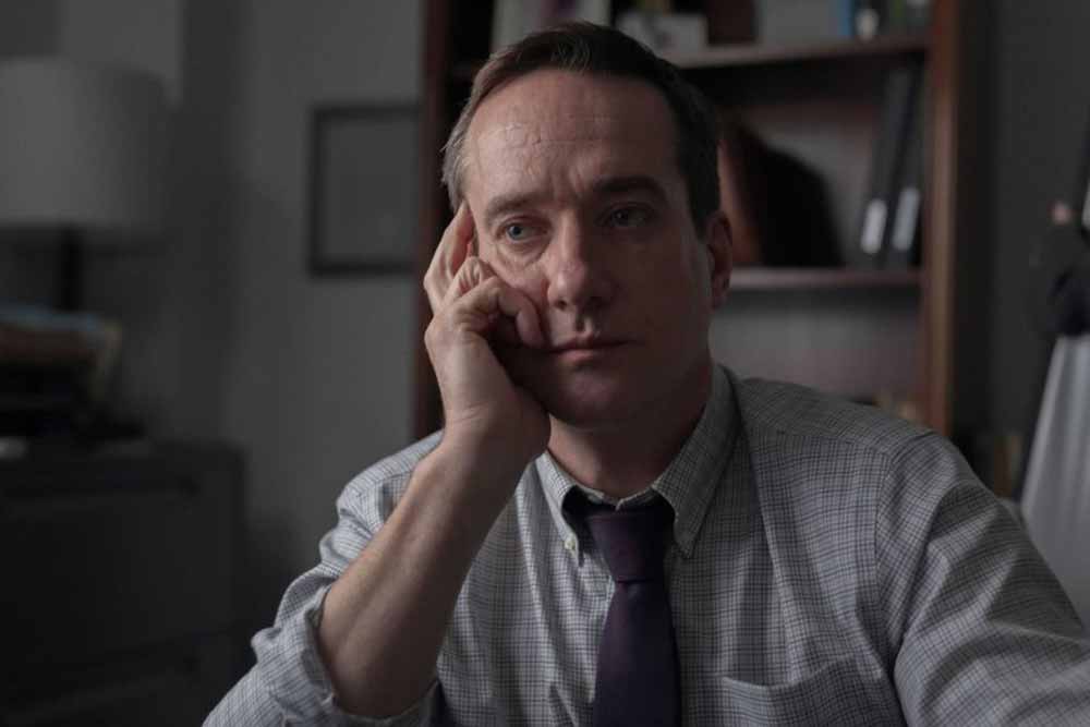 Macfadyen sits at a desk, his head resting against his hand as he listens to Jane speak out of frame.