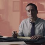 A still of Matthew Macfadyen sitting at a desk in The Assistant, set against a pale pink background.