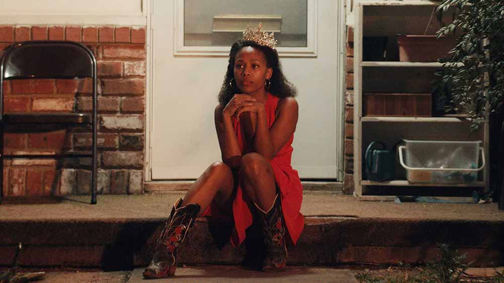 Nicole Beharie sits on a porch in a red dress, wearing a tiara, in Miss Juneteenth.