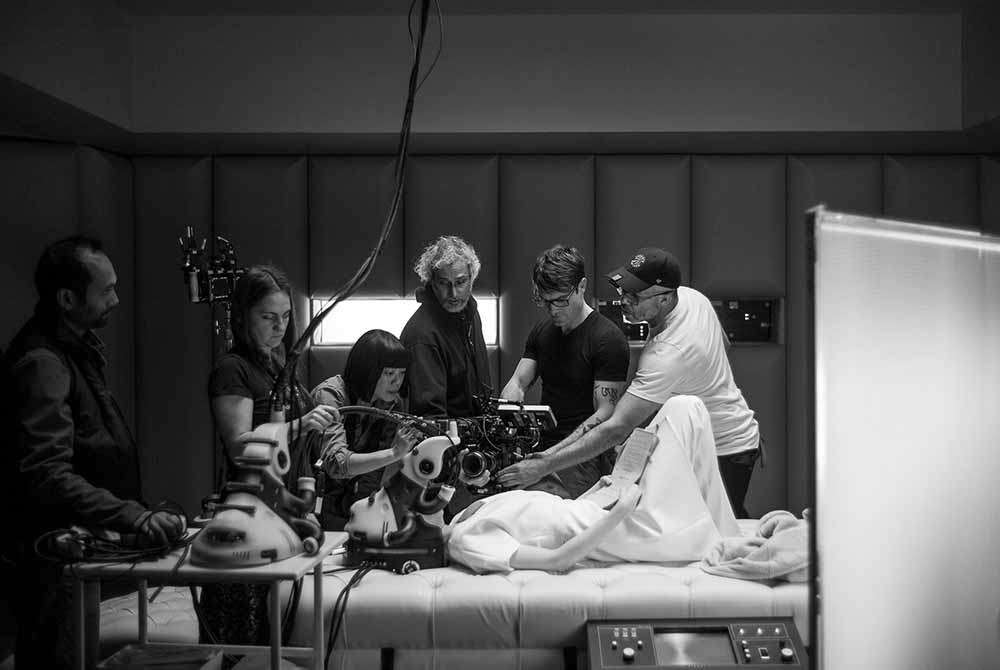 Several members of the filmmaking crew gather around Andrea Riseborough's character as she lies down on what looks like a surgical table. The still is in black and white.