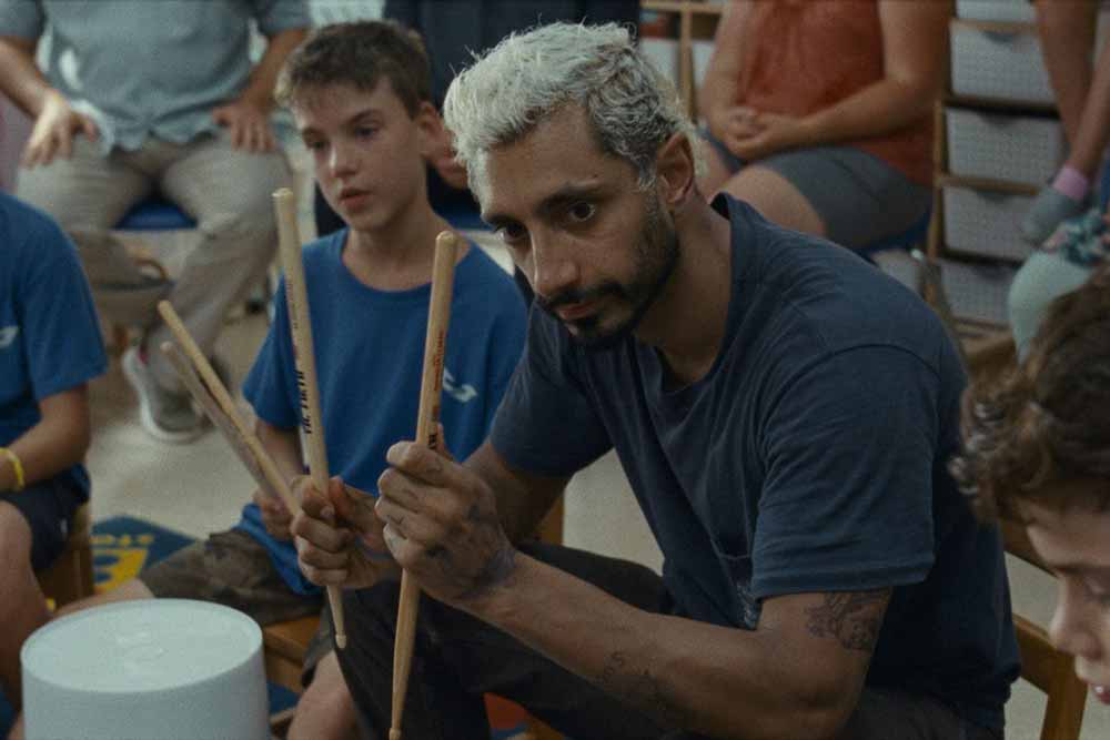 Riz Ahmed holds up two drumsticks to show a group of young children in Sound of Metal.