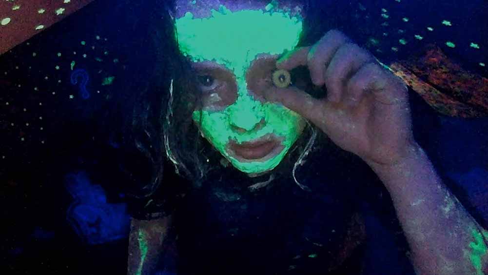 A still of Anna Cobb, one of the most exciting emerging actors at Sundance 2021, in We're All Going to the World's Fair. She is holding a small glass eyeball to her own eye and wearing glow in the dark face paint.