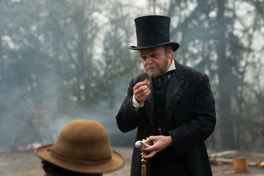 A man in a top hat breathes in the smell of a small cake in First Cow.