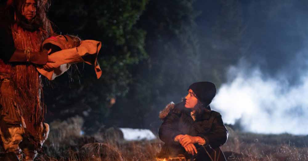 A young woman sits by a campfire at night, and is approached by a man from the left of frame, in Monkey Beach.
