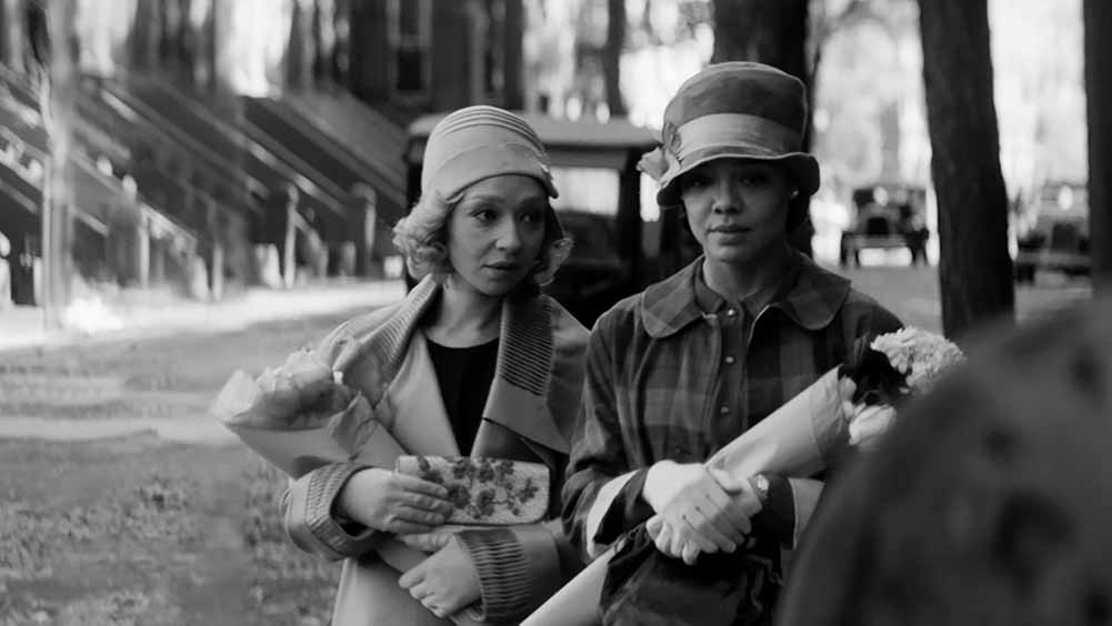 In this black and white still from Passing, two women walk down the street together in hats and heavy coats, holding a bundle of flowers each.