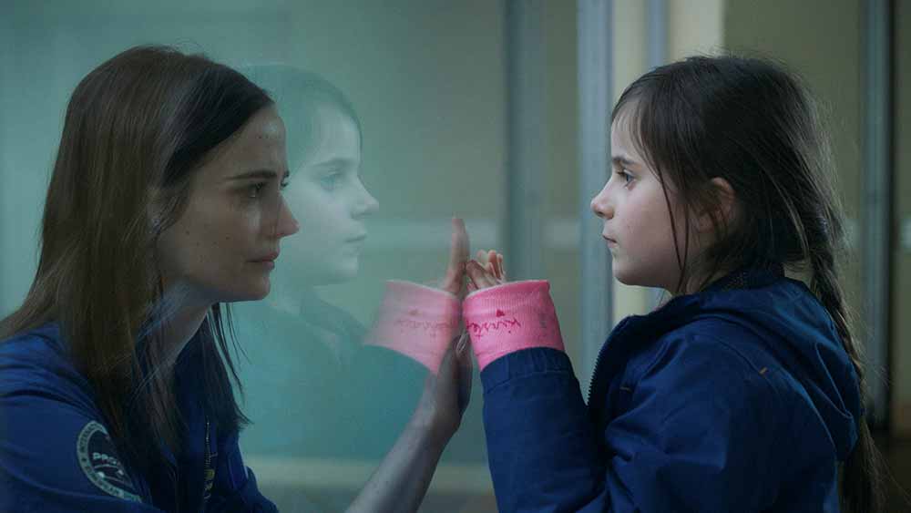 A mother and daughter stand on opposite sides of a pane of glass, their hands up to the glass, overlapping each other. This still is from Proxima.
