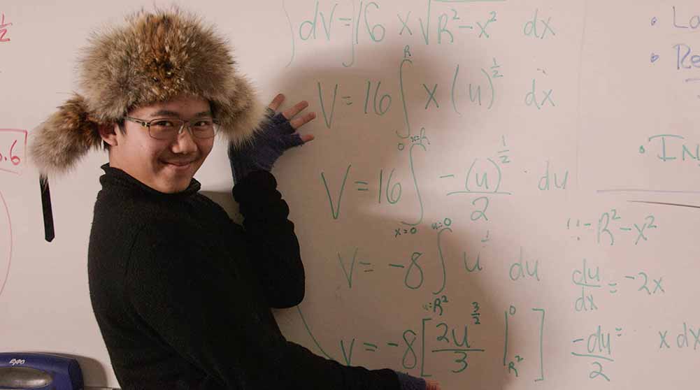 A student in a fluffy hat stands in front of a whiteboard full of math equations.