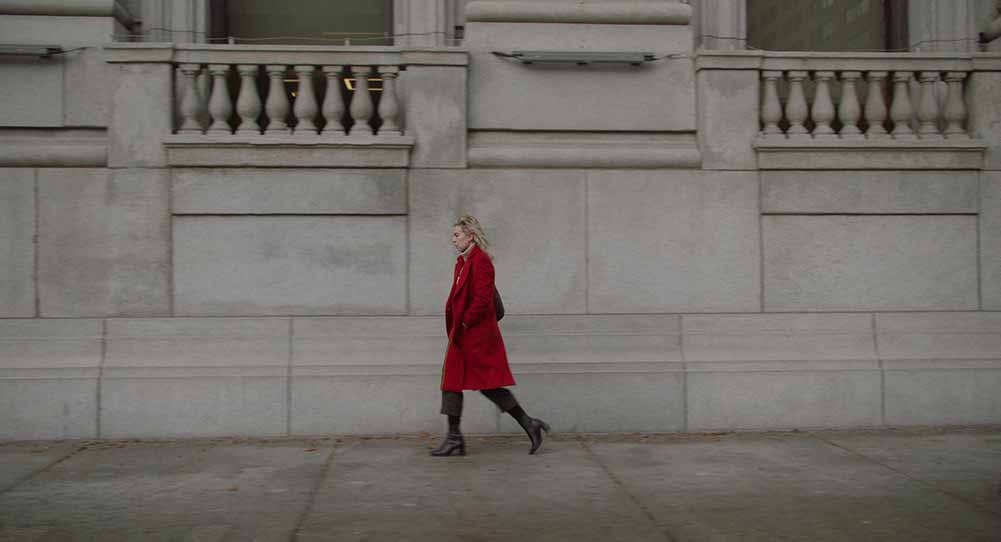 An extreme wide shot of Vanessa Kirby in a bright red coat walking along the pavement in Boston.