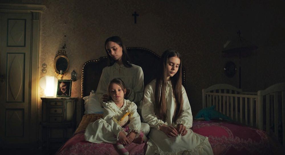Three sisters sit together on a red velvet bed, wearing white nightgowns, in Buio, which screened at Final Girls Berlin 2021.