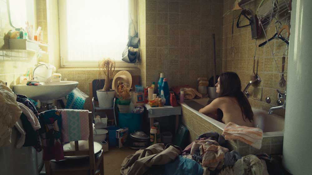 A naked woman sits in a bathtub, surrounded by clutter, in Time of Moulting.