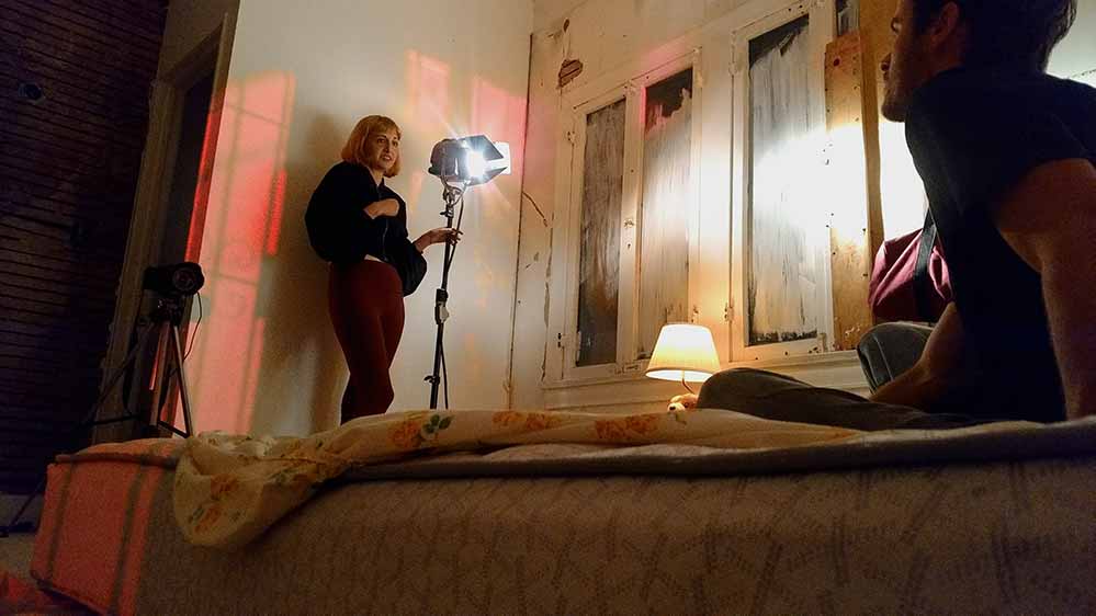 Gillian sets up a large film light in front of a man on a bed who she plans to murder.