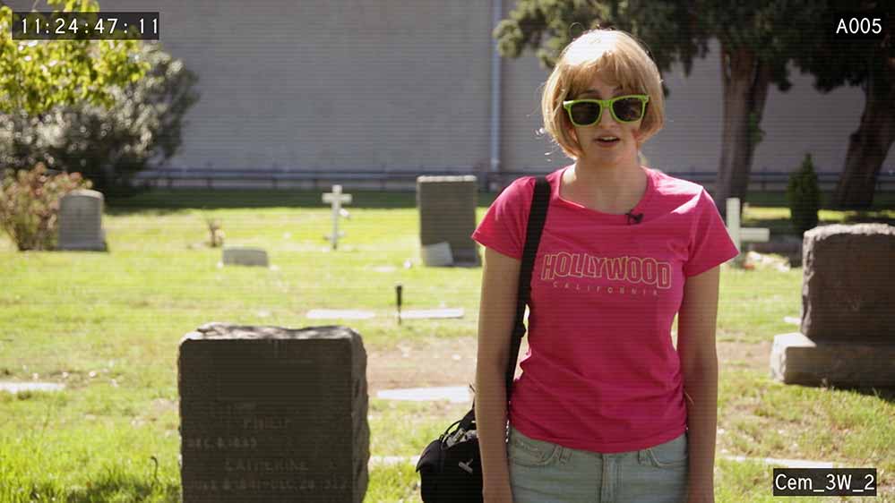 A still of Gillian Wallace Horvat standing in a cemetery, wearing sunglasses and a garish pink shirt, in I Blame Society.