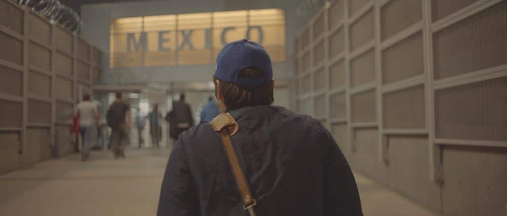 A shot of a young man's back as he walks toward a border sign that reads 'Mexico'. A still from Fernanda Valadez and Astrid Rondero's Identifying Features.
