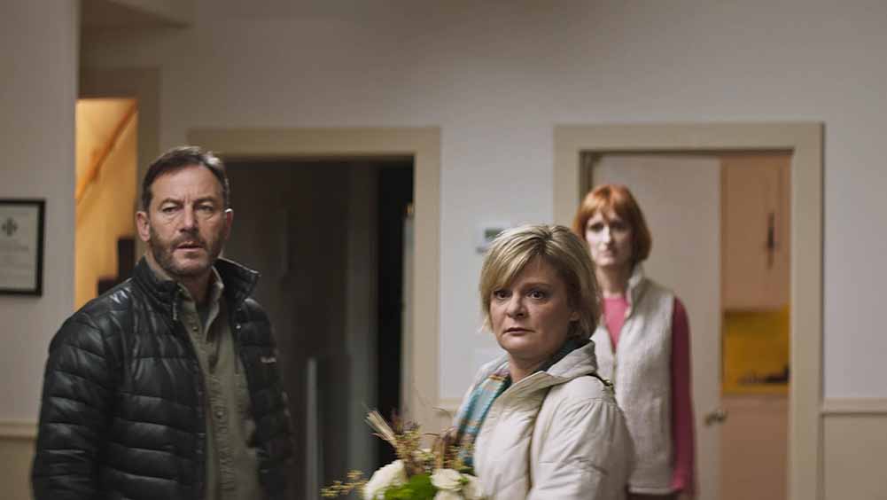 A still from Mass, of three people standing in a room, one of them being Martha Plimpton.