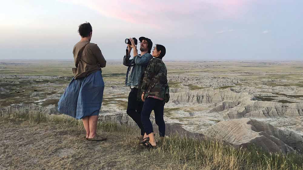 Frances McDormand, Joshua James Richards, and Chloé Zhao stand atop a hill, a vast and rocky landscape behind them.