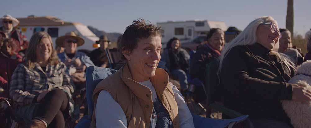 A group of older Americans, Frances McDormand amongst them, sit on camper chairs near their trailers, smiling together.