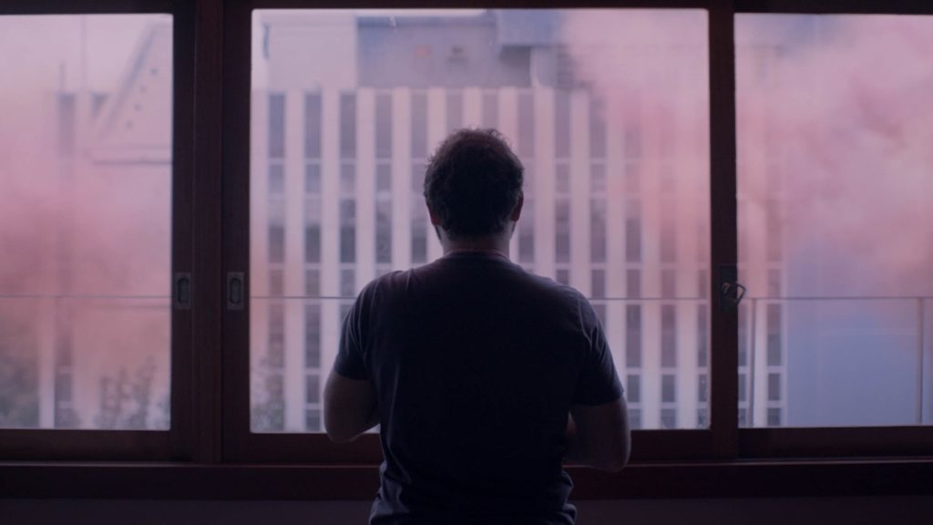 Eduardo Mendonça stars as Yago in The Pink Cloud. Yago is seen in near silhouette from behind, staring out the window at the pink cloud.
