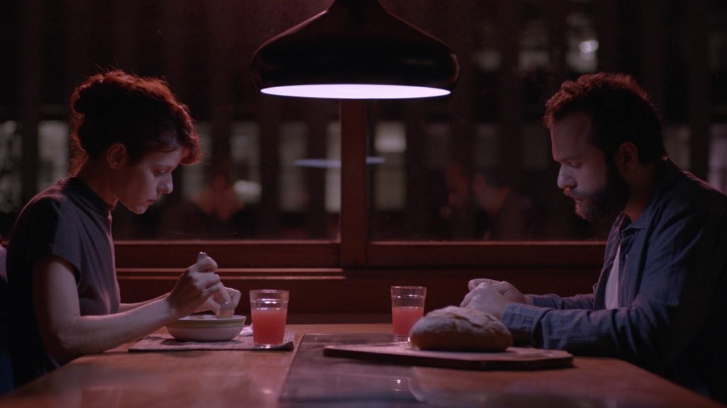 Giovana (Renata de Lélis) and Yago (Eduardo Mendonça) share a meal in The Pink Cloud. The scene is bathed in the pink light of the cloud.