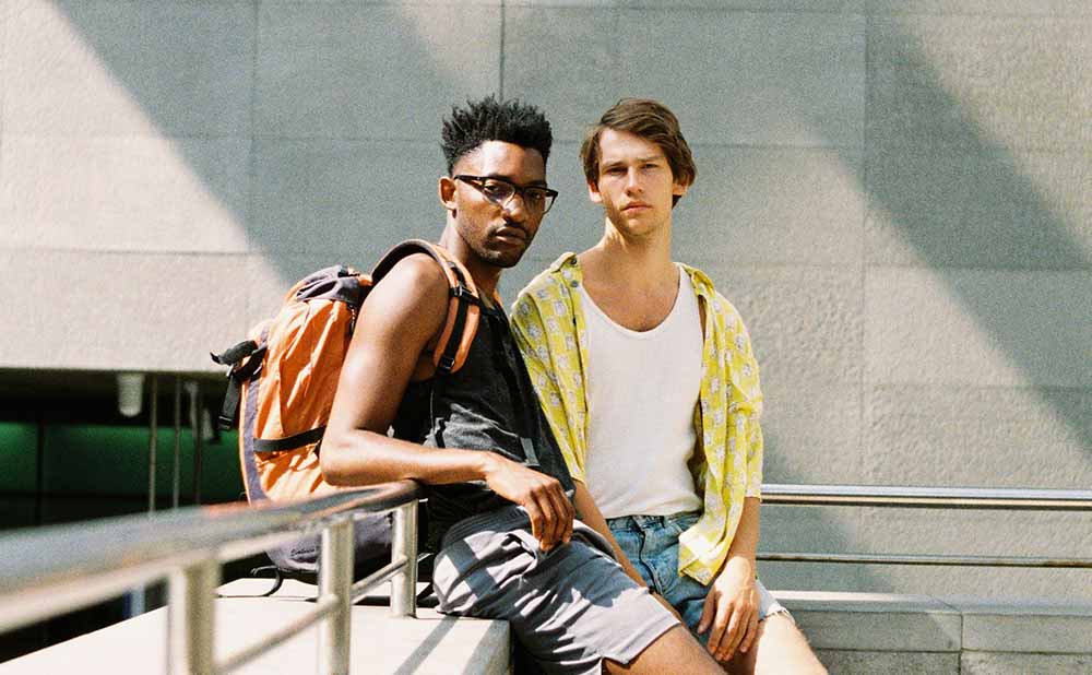 Two young men lean against an outdoor banister in the summer sun in Boy Meets Boy, a BFI Flare 2021 film.