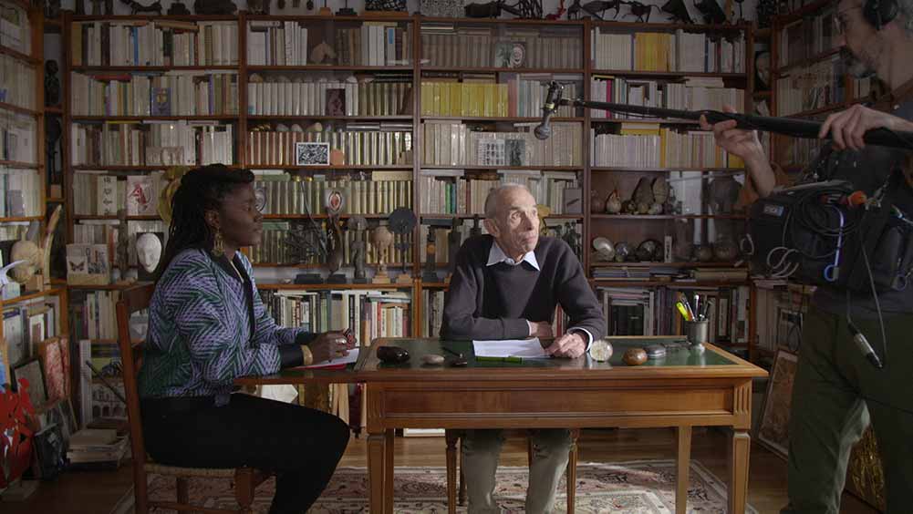 Alice Diop sits at a table with the elderly Pierre Bergounioux, a wall of books behind them.