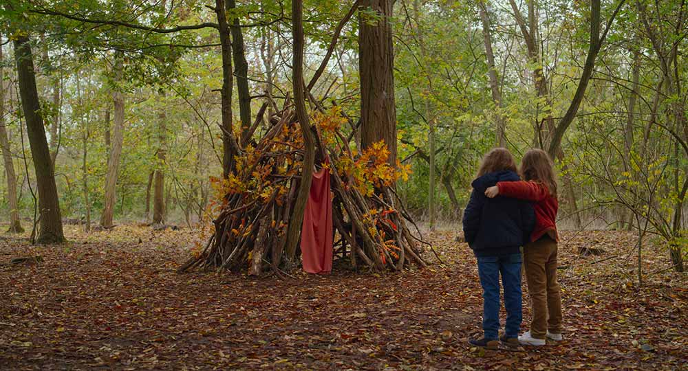 Two young girls stand side by side, arm in arm, looking on at a tree fort they made, in Petite Maman, one of the best films of Berlinale 2021.