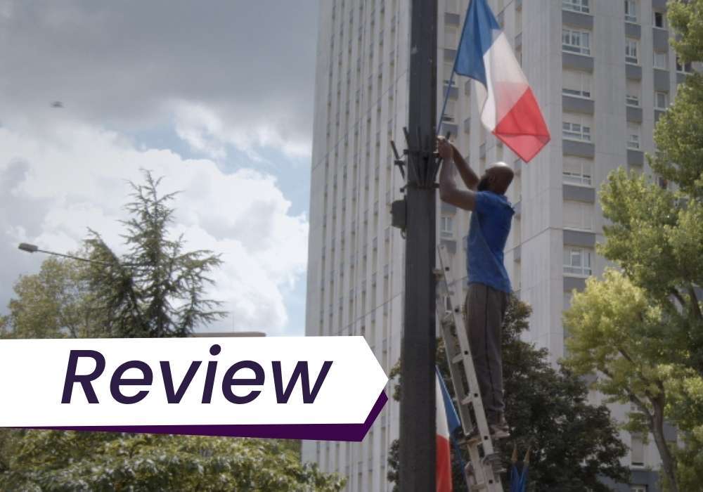 A man hangs up a French flag on an outdoor pole in Nous. The text on the image reads, 'Review'.