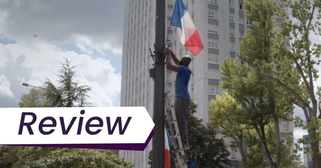 A man hangs up a French flag on an outdoor pole in Nous. The text on the image reads, 'Review'.